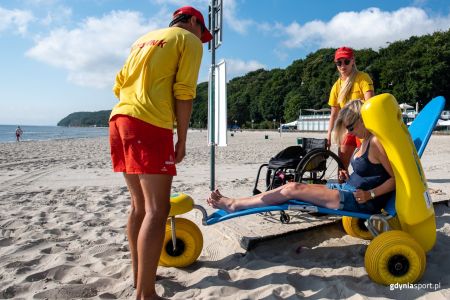 dwójka ratowników stojących na plaży wraz z kobietą siedzącą na wózku kąpielowym