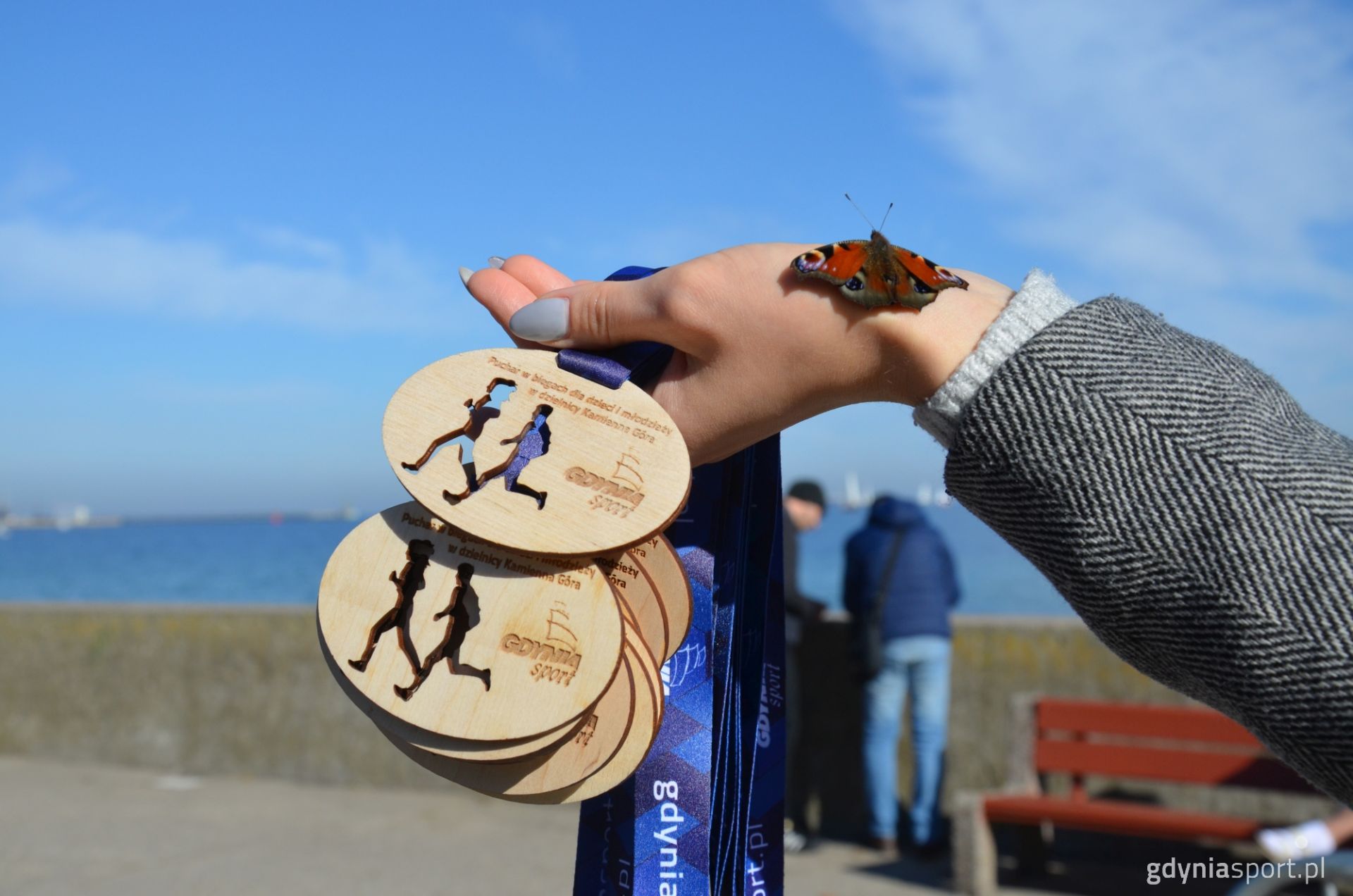 medale trzymane przez kobiecą dłoń z siedzącym motylem na tle morza 